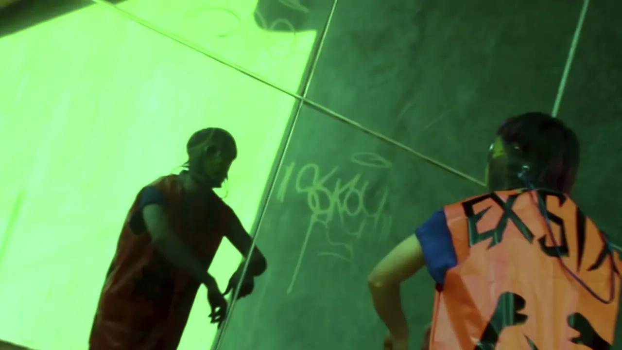 Dos figuras contra un muro verde, una lleva el chaleco naranja EXSIX